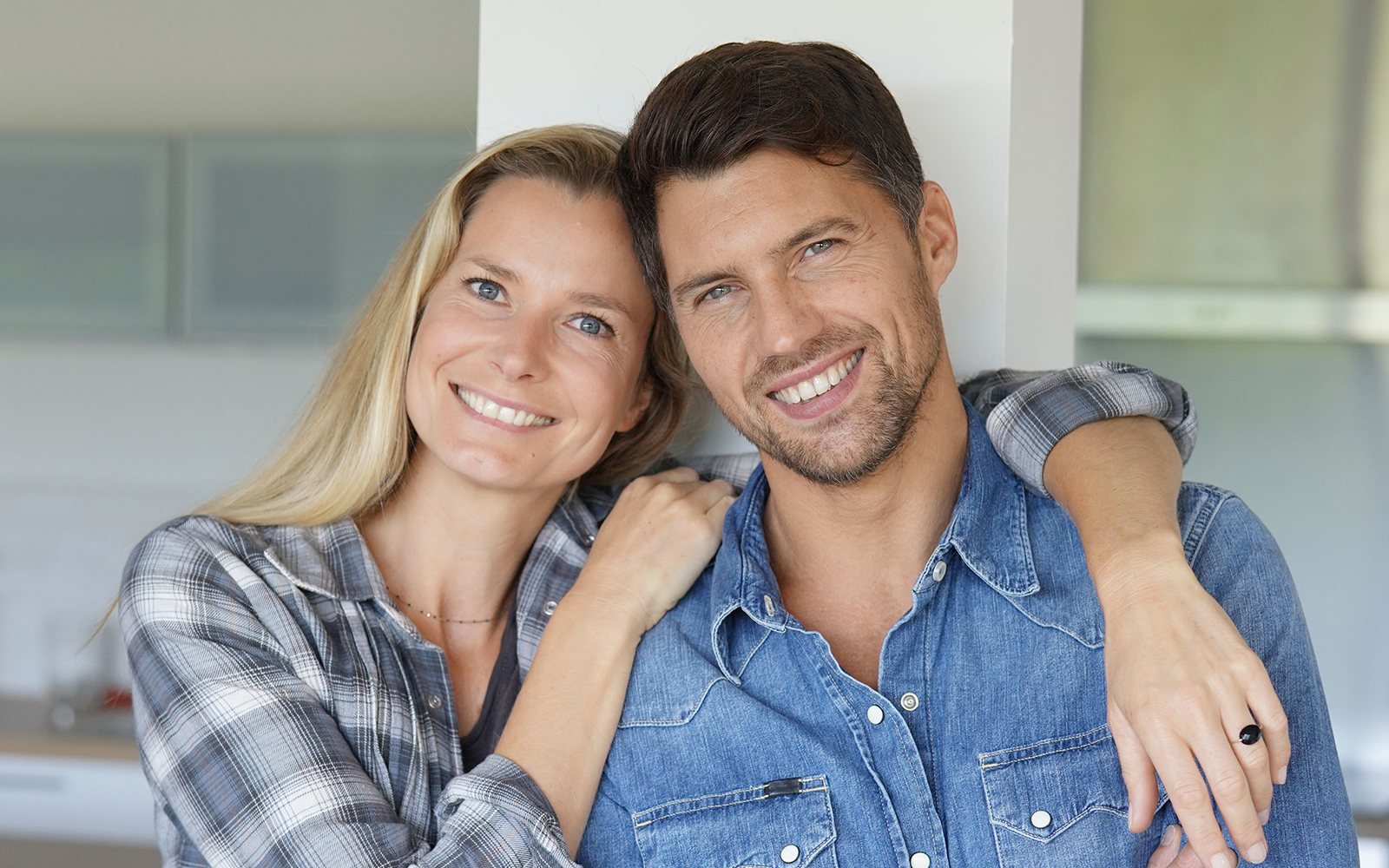 A man and woman are posing together, with the man wearing a plaid shirt and the woman in a denim jacket, both smiling and embracing each other against a white background.