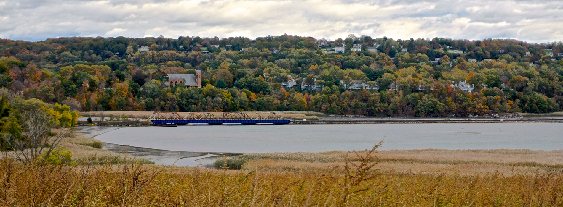 The image shows a scenic view of a river with a barge passing by, surrounded by autumn foliage on trees, and a distant shore with houses and a boat dock.