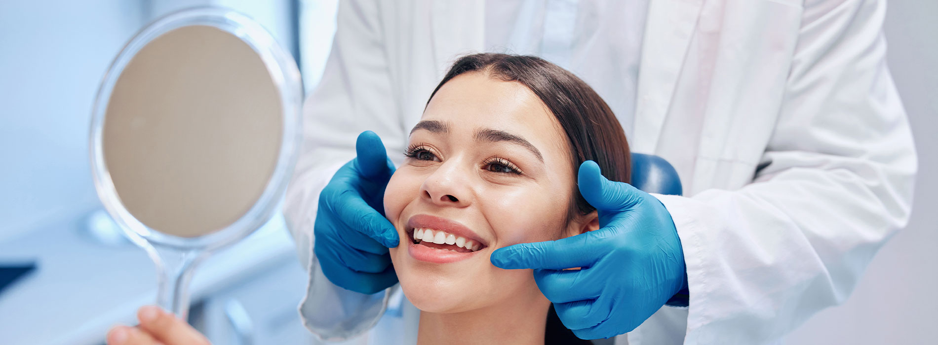 The image shows a person sitting in a dental chair with a smile on their face, receiving dental care from a professional wearing blue gloves and a white coat, with a mirror reflecting the scene.