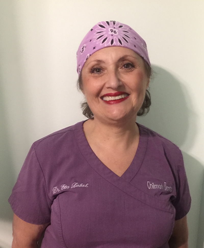 The image shows a person wearing a purple headband, a white shirt with a name tag, and scrubs, posing for a portrait.