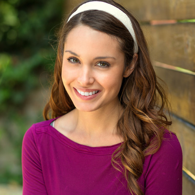 The image shows a woman with long brown hair wearing a purple top and a white headband, smiling at the camera.