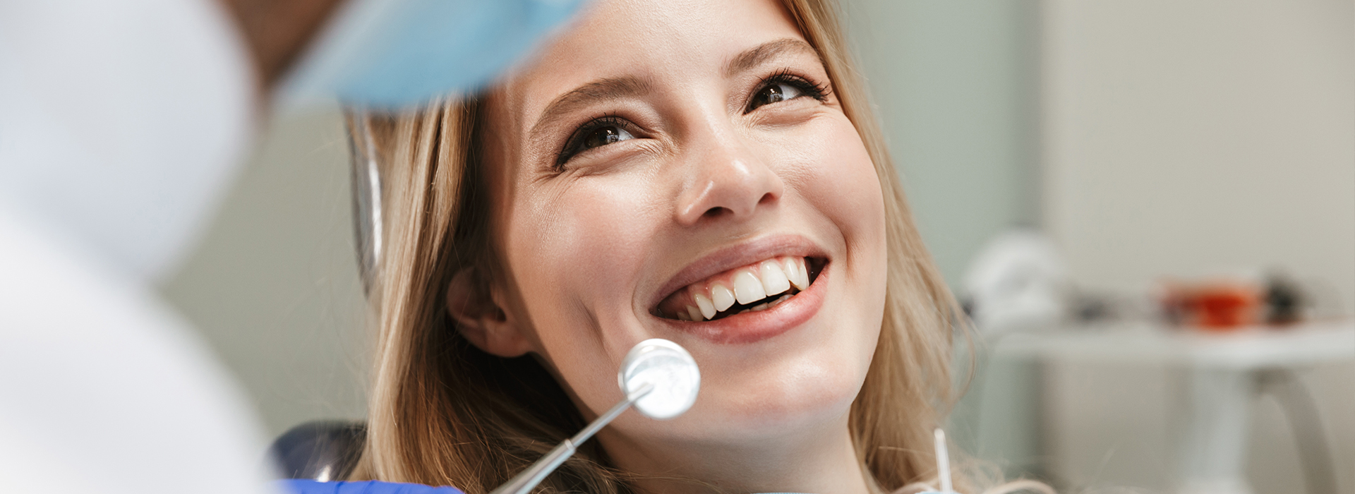 The image shows a woman with blonde hair sitting in a dental chair, smiling at the camera, undergoing a dental examination with a dentist visible behind her, wearing a white coat and stethoscope.