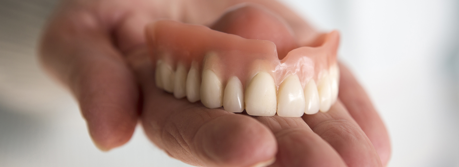 The image shows a person holding a denture with teeth against a blurred background, featuring an older individual s hand displaying a set of artificial teeth.