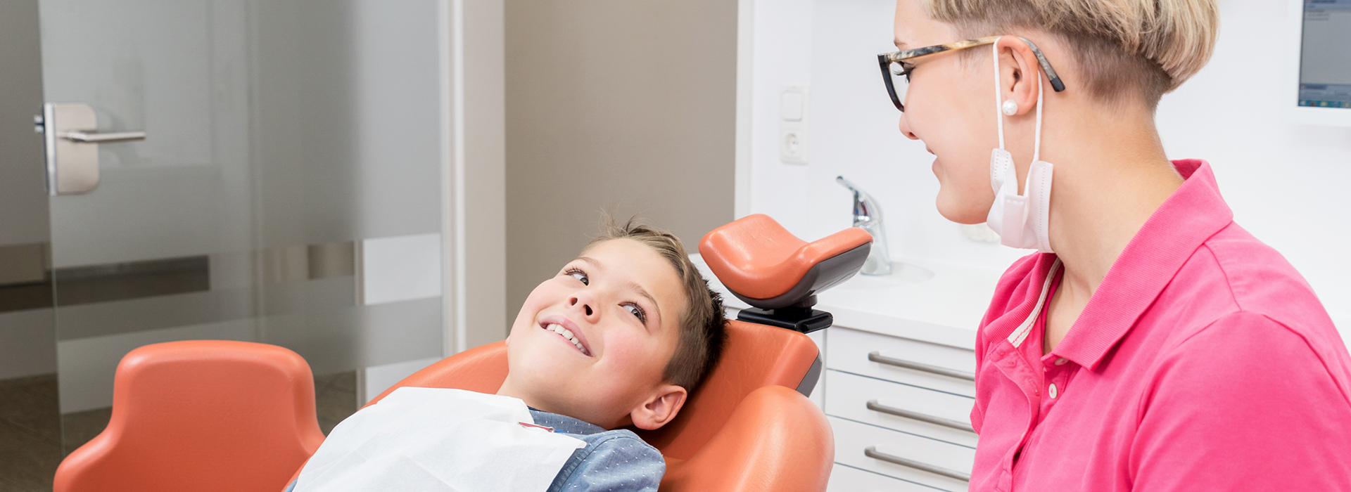 A woman sitting at a desk with a young boy standing next to her, both smiling, looking towards the camera.