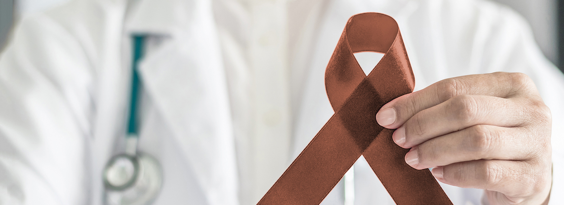 The image shows a healthcare professional holding up a red ribbon with a white cross symbolizing medical aid, against a background that includes a blurred figure of a person and what appears to be a hospital setting.