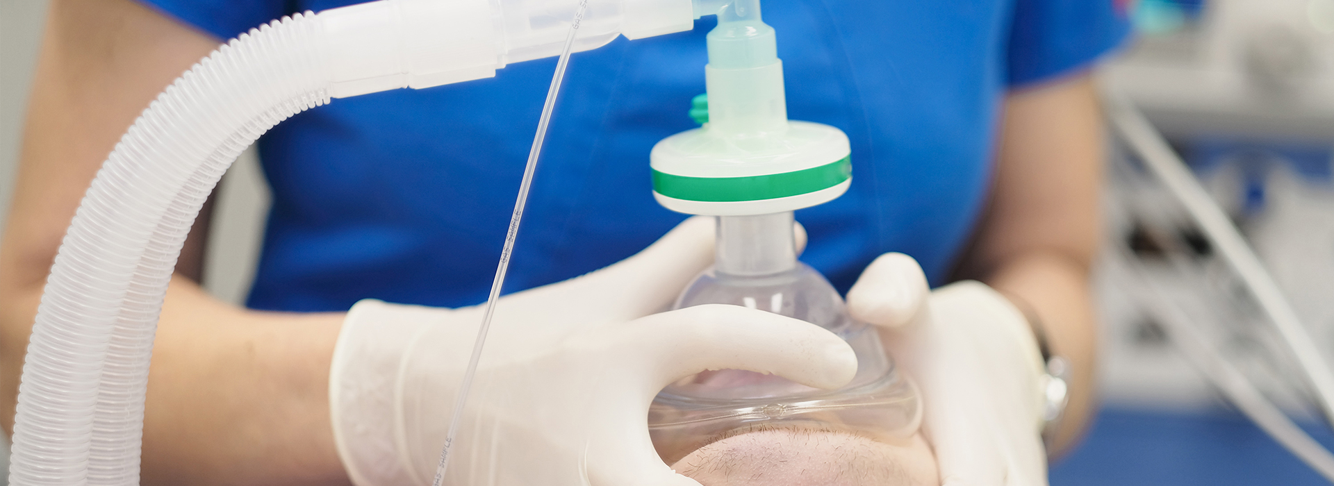 The image shows a medical setting with a person wearing white gloves holding a transparent plastic bottle containing a liquid substance, which appears to be a medication or solution, against a background that includes medical equipment and supplies, suggesting a healthcare professional in the process of administering or preparing for administration of the substance.