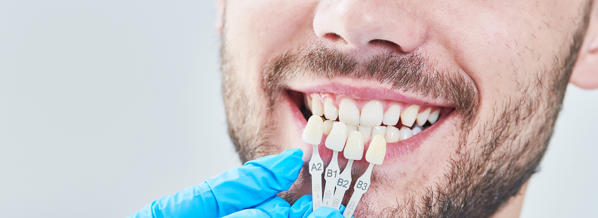 The image shows a person with a toothy smile holding a toothbrush with toothpaste, wearing a blue surgical mask and white gloves, while being photographed from below against a blurred background.