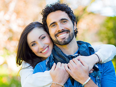 A man and woman embracing each other with smiles, set against a blurred outdoor background.