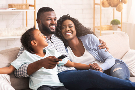 The image depicts a family of four sitting on a couch, with the father holding a remote control and smiling at the camera, while the mother embraces him from behind.