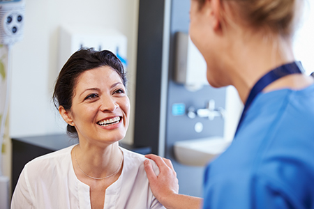The image depicts two individuals in a medical setting a woman with a smile, wearing a white top, stands next to a person who appears to be a healthcare professional in scrubs, engaging in conversation.