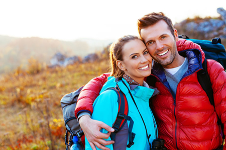 A man and woman are posing for a photo outdoors, with the man wearing a red jacket, both holding each other, and they appear to be on a hiking trip during sunset.