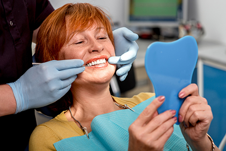 The image shows a woman sitting in a dental chair with a large smile, holding up a blue model of a tooth, while a dental hygienist attends to her with tools such as a mirror and dental pick.