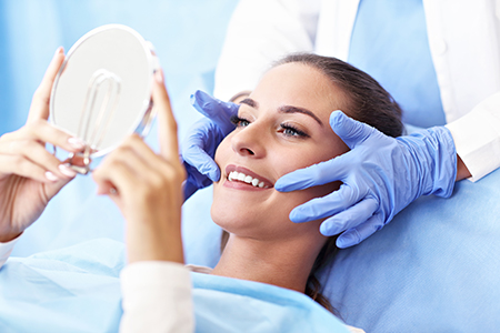 The image shows a person seated on a dental chair with a large mirror placed over their face, being attended to by two individuals wearing medical gloves and a blue gown, who appear to be dental professionals performing an examination or treatment.