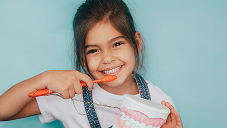 A young girl brushing her teeth with an electric toothbrush while holding a toothpaste tube.
