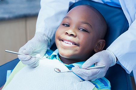 The image shows a young boy seated in a dental chair, smiling at the camera while wearing a dental mask with a mouthguard, receiving dental care from a dentist who is adjusting his mouthpiece.