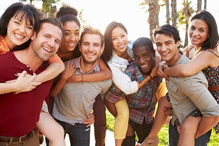 This is a color photograph featuring a group of young adults posing together for a picture with smiles on their faces. They are standing close to each other, embracing some members of the group, under bright daylight, possibly at an outdoor venue.