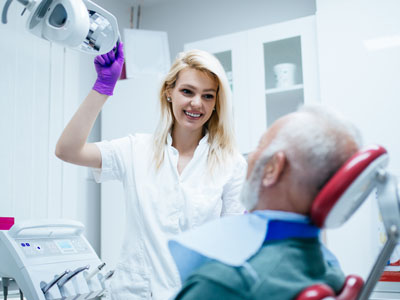 The image shows a woman standing next to an older man who is seated in a dentist s chair, receiving dental care  she is holding a mirror up to his face.