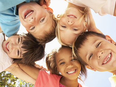 The image shows a group of children posing together with smiles on their faces, under a bright sky.