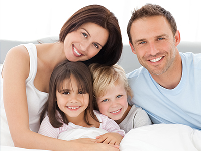 A family of four adults and three children lying together on a bed, smiling at the camera with a happy expression.