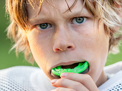 Two images of a young person with blonde hair and blue eyes, holding a green object close to their mouth, which appears to be a toothbrush or similar item. The individual has a scowling expression and is wearing a sports jersey.