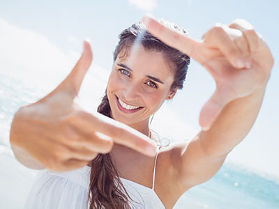 The image shows a woman taking a selfie with her hand held up to her face, framing the shot. She appears to be at the beach, with clear skies and bright sunlight visible in the background.